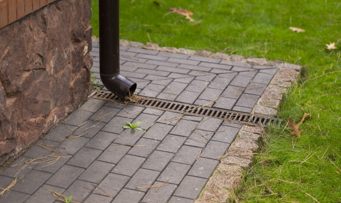 An exterior drainpipe leading to an outside strip drain surrounded by pavers and grass.